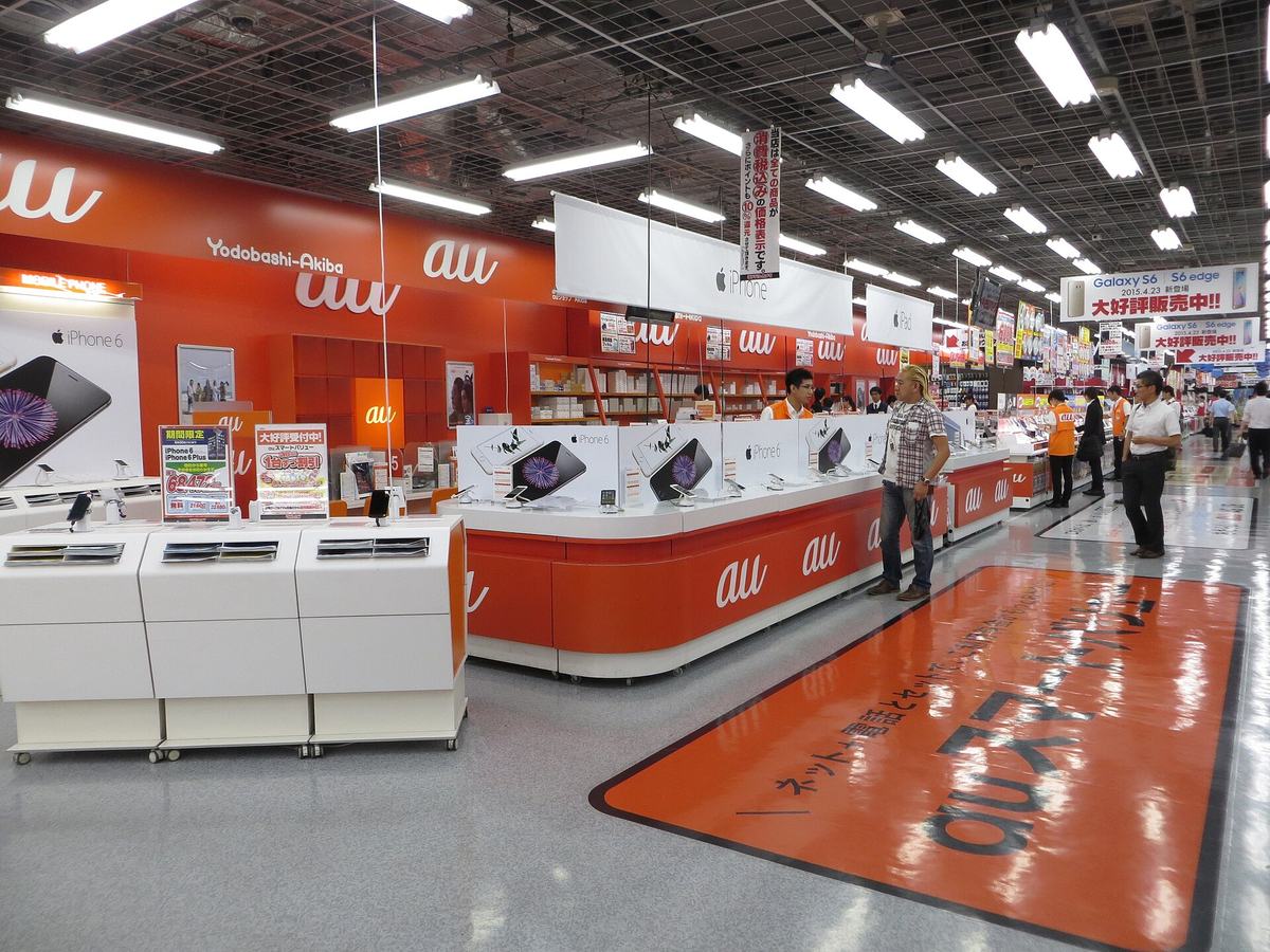 家電量販店内の赤いauコーナー。auロゴが並ぶカウンターで店員が接客している