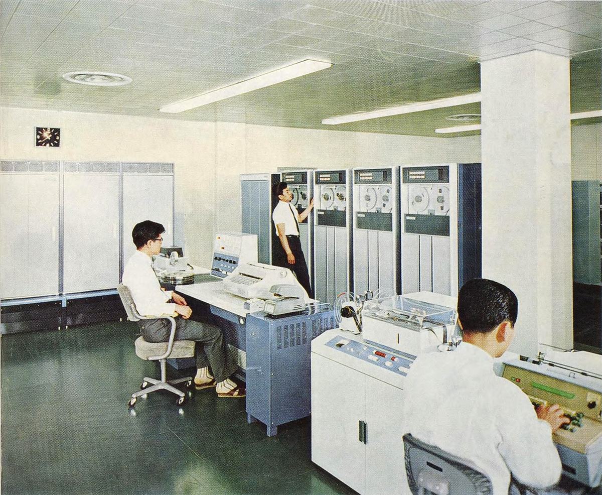 1970年前後の計算機室。磁気テープ装置が並び、白衣の技術者が操作している